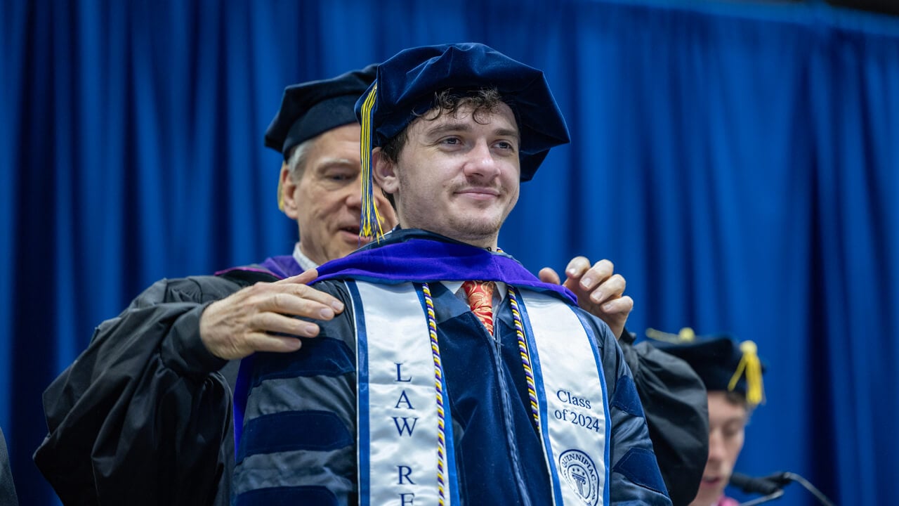 A professor places a doctoral hood on a graduate who wears a white stole that reads Class of 2024