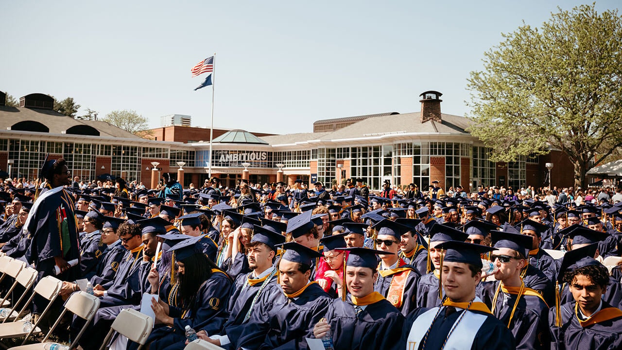 A wide angle photo of graduates sitting in their chairs on the quad.